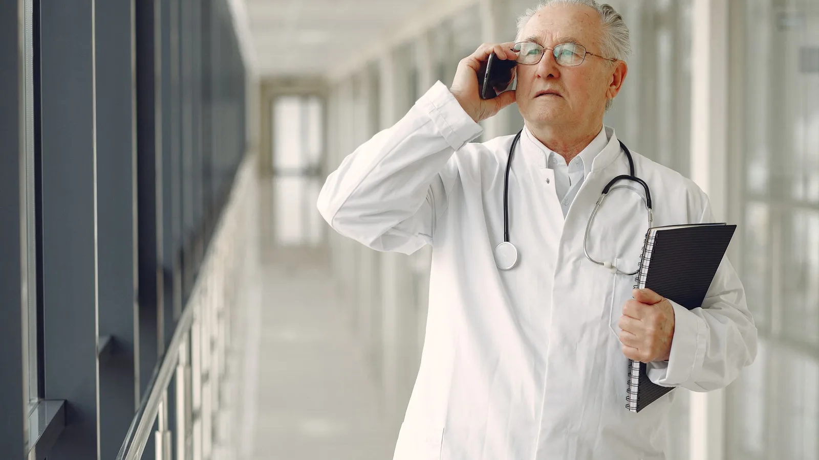 Serious mature male doctor in medical uniform standing with stethoscope and notebook while talking on smartphone in clinic corridor