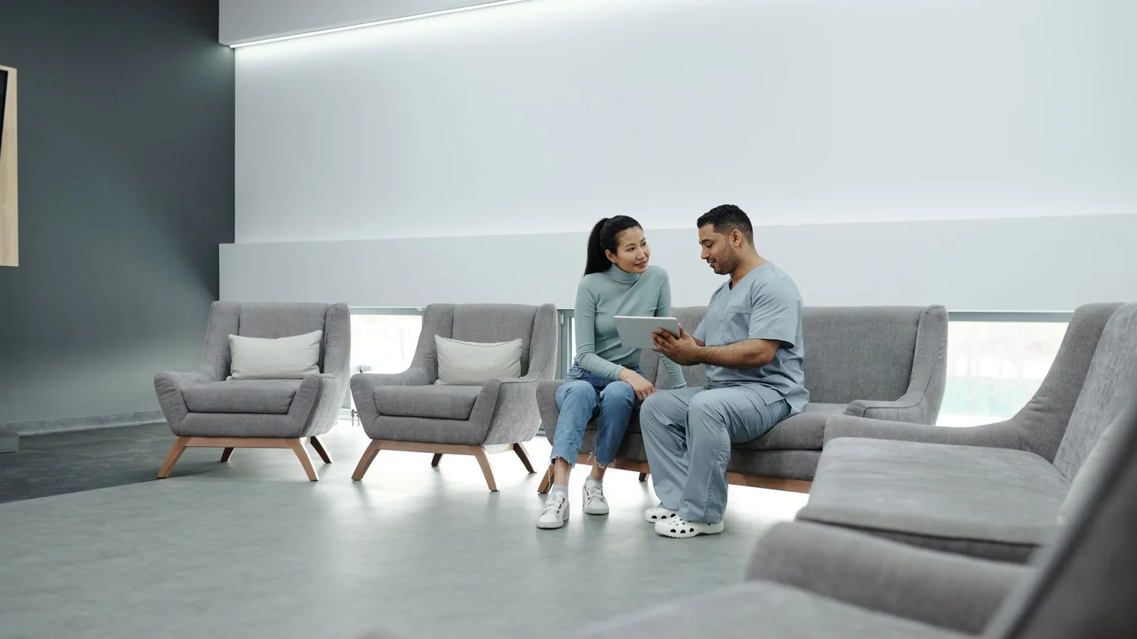 A doctor in scrubs consulting a female patient in a modern clinic waiting room, using a tablet.